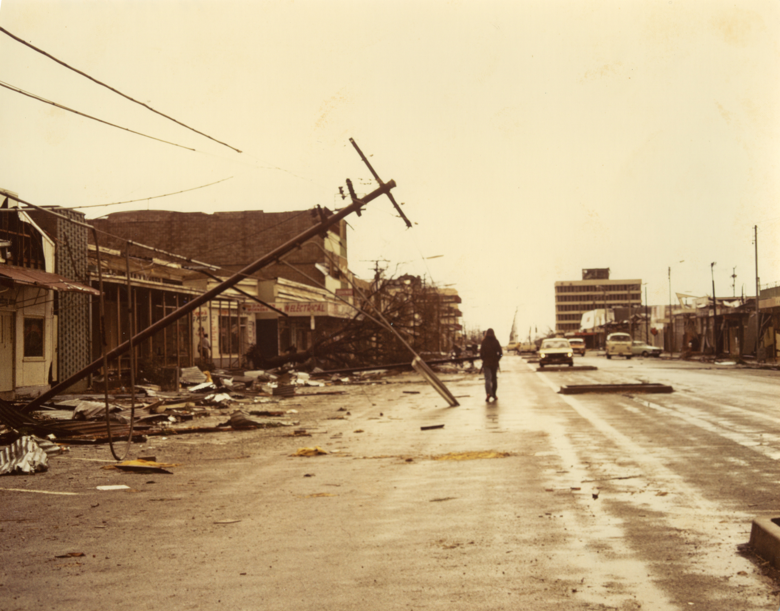 Person walking down a street in Darwin after Cyclone Tracy, surrounded by severely damaged buildings and debris.