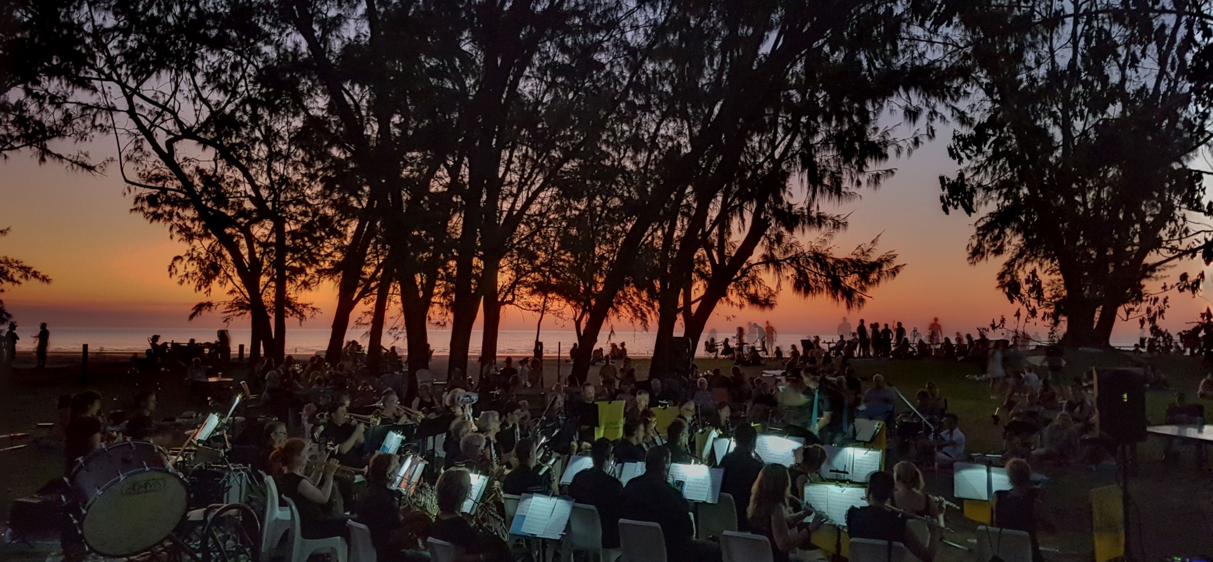 Arafura Wind Ensemble performing on the grass at Casuarina Surf Club, with the sun setting behind trees and a view of the ocean.